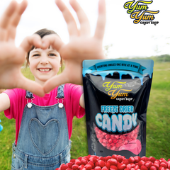 Child making a heart shape with hands, Yum Yum Candy Shop freeze-dried candy packaging, and red candy on grass.