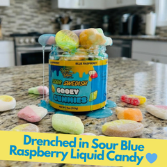 The gummy jar sitting on a granite kitchen countertop. Several loose gummy candies are scattered around the base of the jar, which is drenched in a bright blue raspberry liquid candy.
